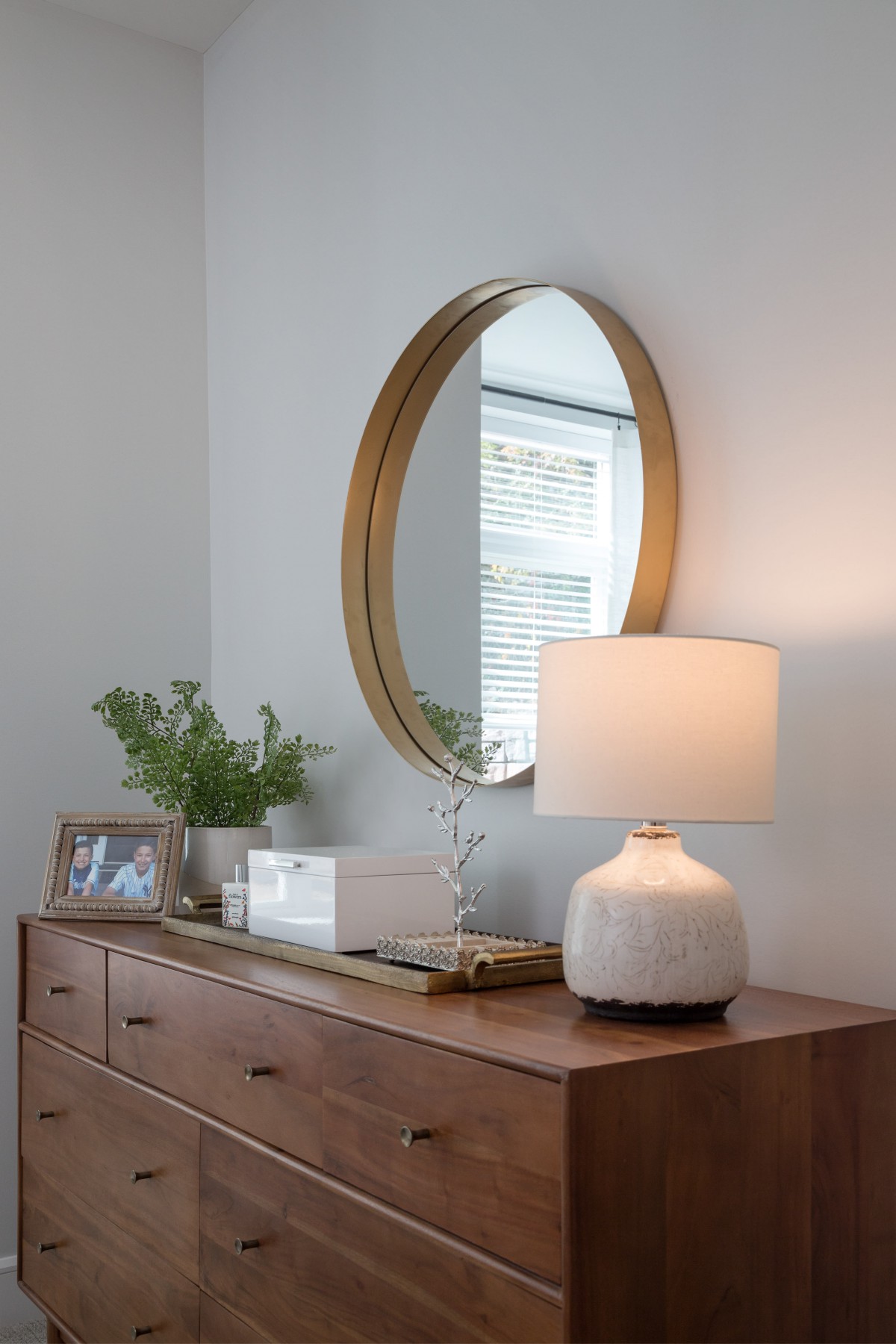 A stylishly decorated dresser featuring a round mirror, modern lamp, framed photo, and greenery.