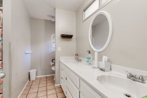A clean and modern bathroom featuring a double vanity with a round mirror, tile flooring, and ample natural light.