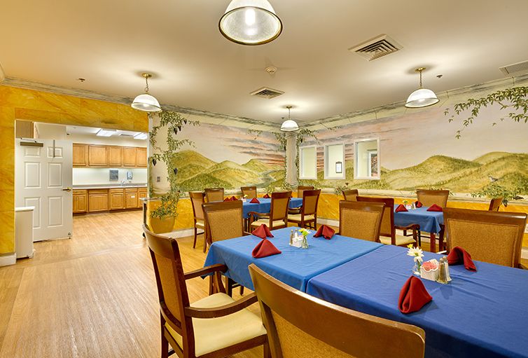 A cozy dining room with blue tablecloths and a mural on the wall, connected to a kitchen area.