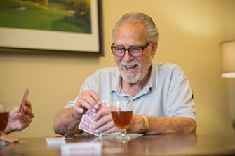 An elderly man with glasses smiles while playing cards at a table with a drink.