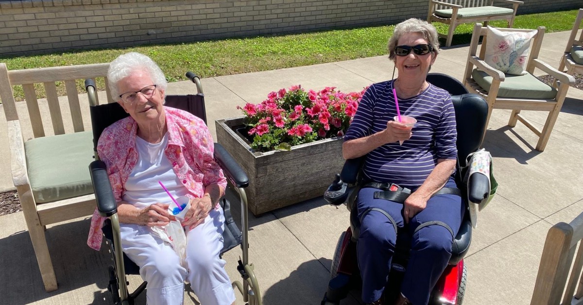 Two women sitting outside in wheelchairs near a flower planter.