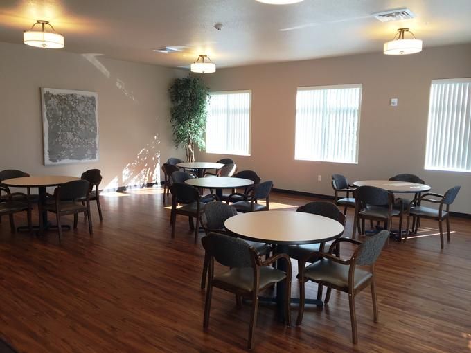 A spacious dining room with round tables and chairs on a wooden floor, lit by ceiling lights and natural light from the windows.