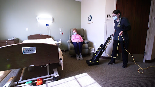 A healthcare worker cleans the hospital room while a patient sits in a chair.