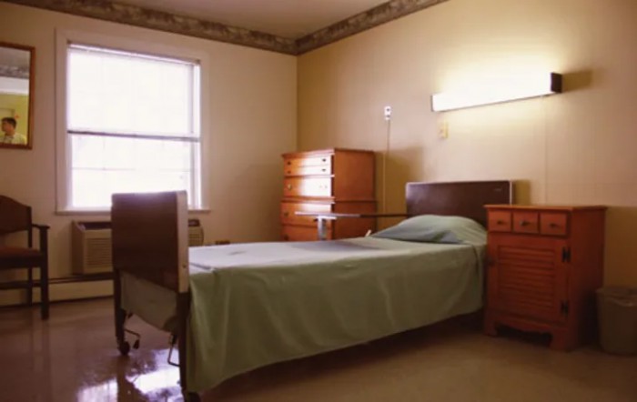 A minimalist hospital room featuring a single bed and wooden furniture, illuminated by natural daylight.
