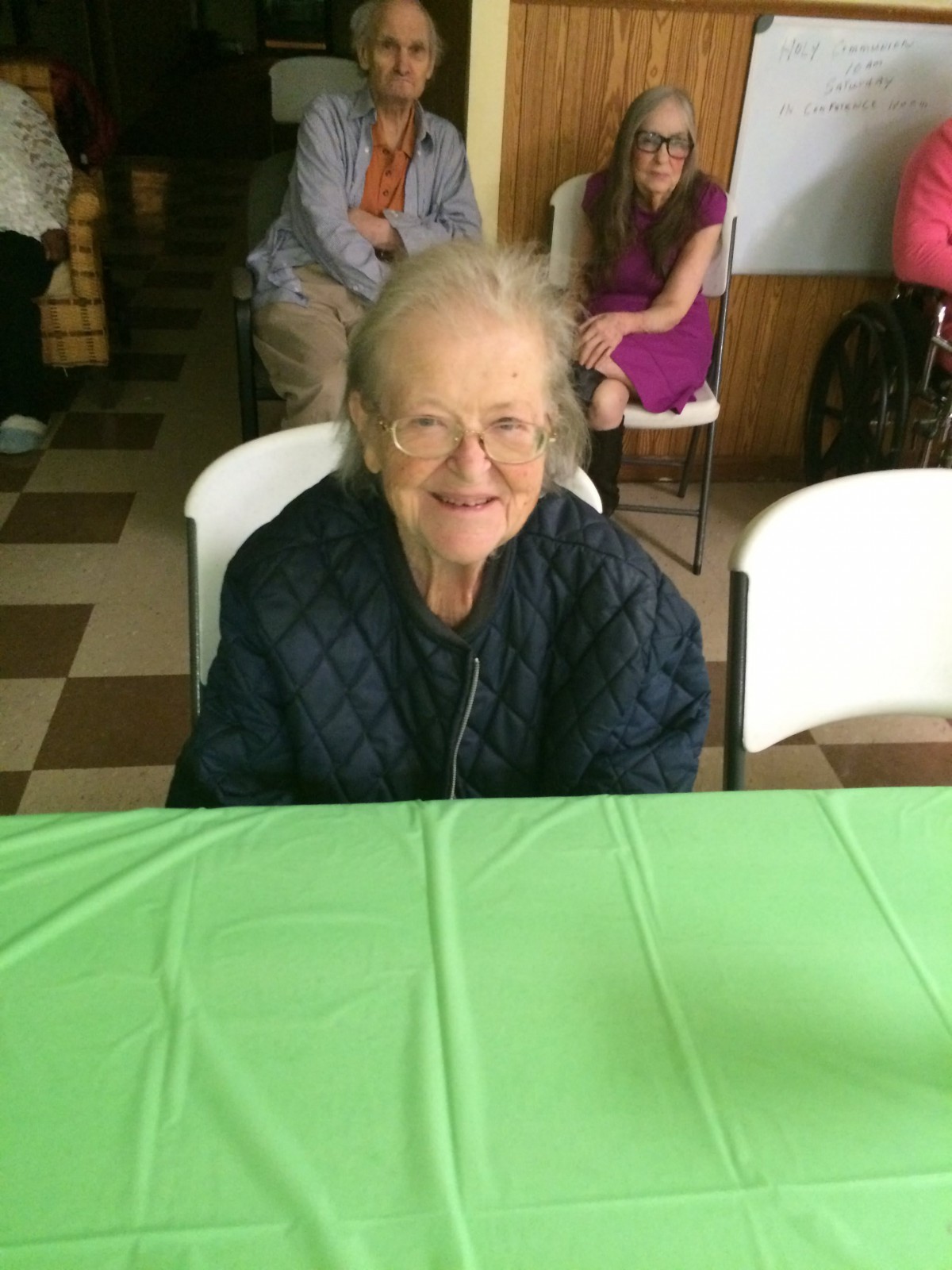 An elderly woman smiling while sitting at a table covered with a green tablecloth.