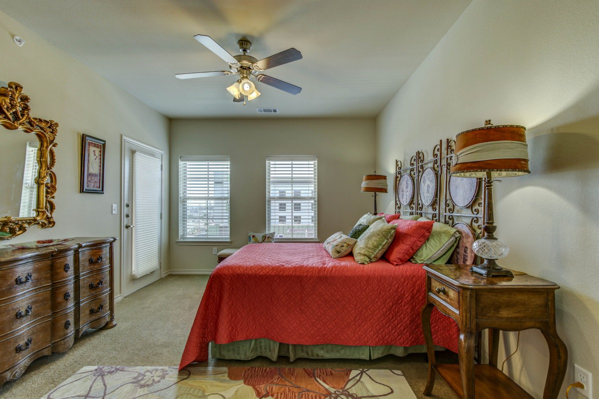 A cozy bedroom featuring a red bedspread, multiple pillows, and a ceiling fan with wooden blades. The room is well-lit by two large windows and is decorated with lamps and matching nightstands.