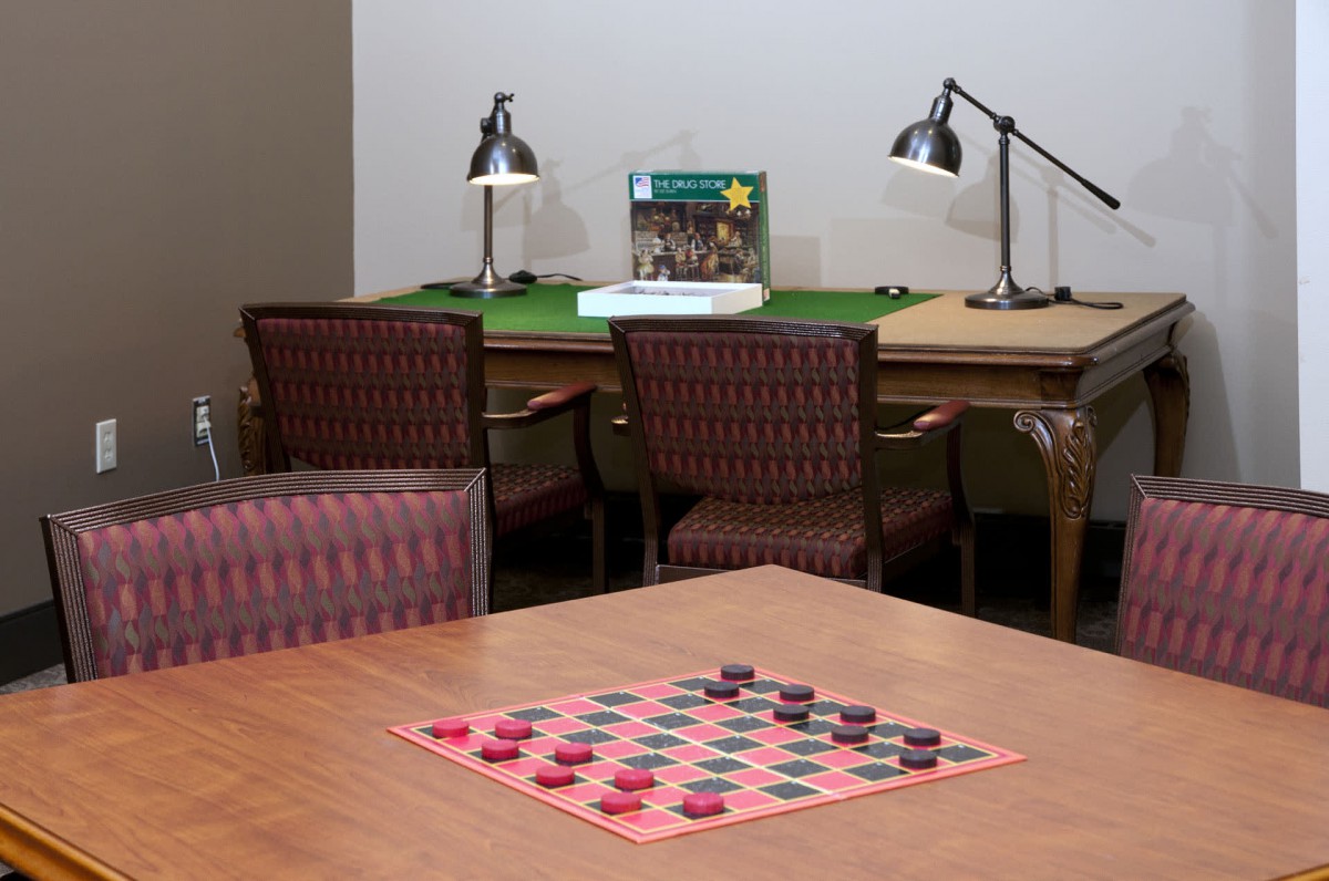 A cozy room with a checkers board set up on a wooden table and two desk lamps in the background.