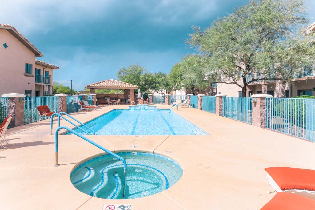 An outdoor swimming pool and hot tub surrounded by a fence, with trees in the background under a bright sky.