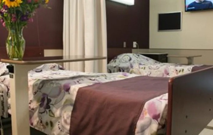A hospital room with two beds, separated by a curtain, featuring floral sheets and a wall-mounted television.