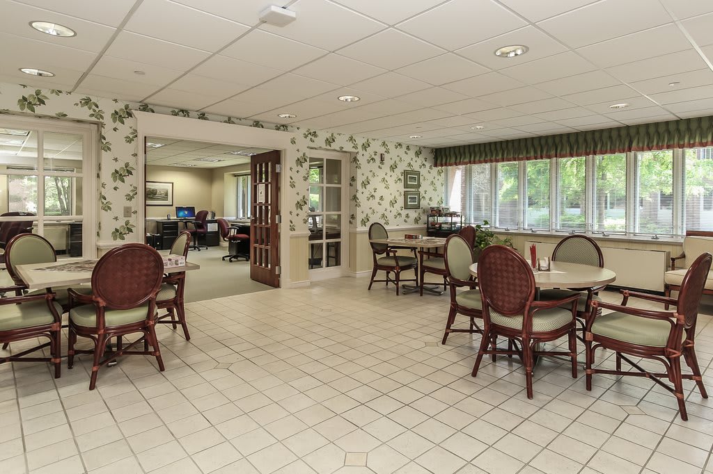 A cozy dining area with floral wallpaper and multiple tables and chairs, featuring large windows and a tiled floor.