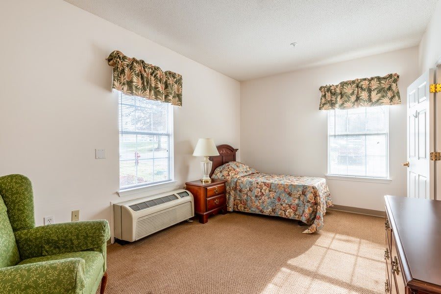 A cozy bedroom featuring a single bed, nightstand with a lamp, windows with floral curtains, and an air conditioning unit.