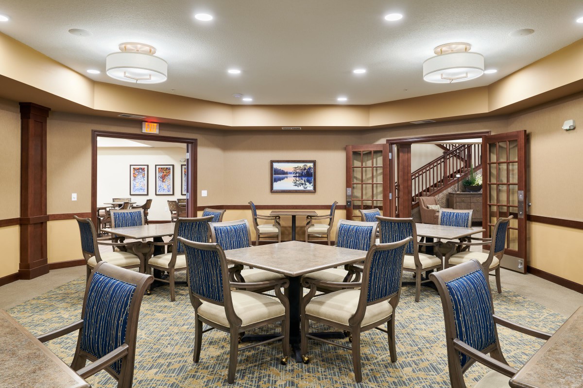 A well-lit dining area with tables and chairs arranged neatly, featuring elegant lighting and a carpeted floor.