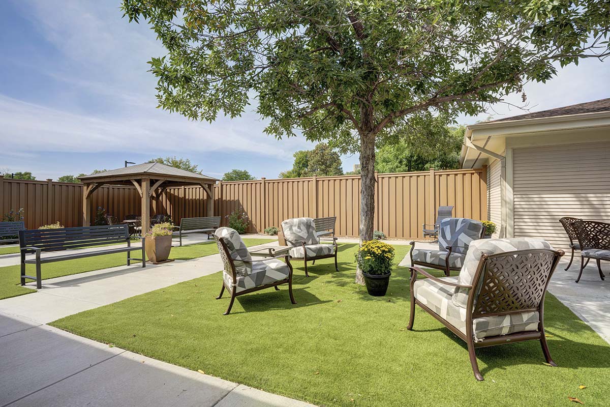 A cozy outdoor seating area in a backyard, featuring chairs around a small table under the shade of a tree.
