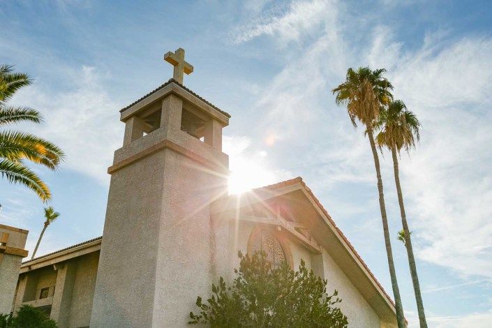 A church with a cross on top, illuminated by sunlight with palm trees nearby.