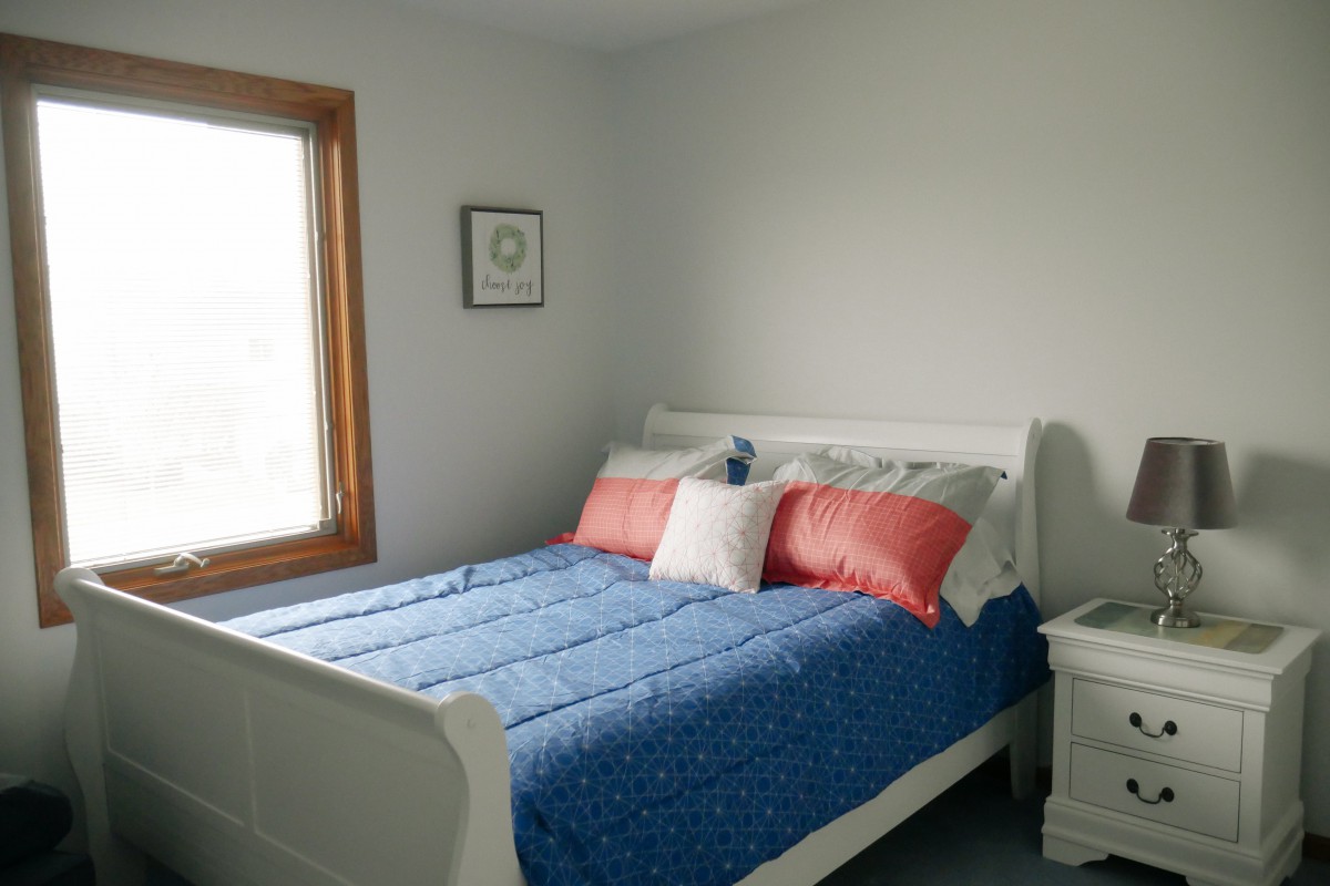 A cozy bedroom with a blue bedspread and colorful pillows, featuring a window and a nightstand with minimalist decor.