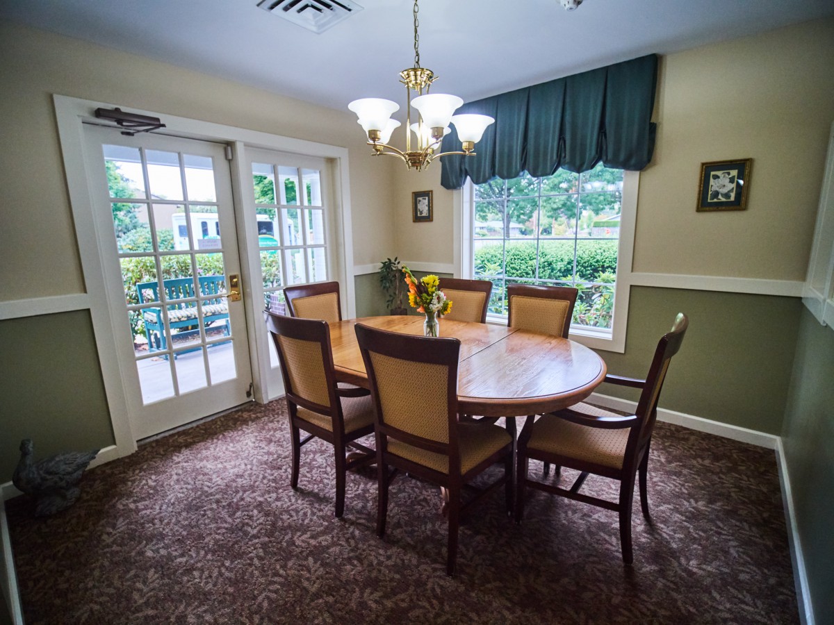 A cozy dining room with a round table and six chairs, illuminated by a chandelier, featuring carpeted flooring and large windows with curtains.