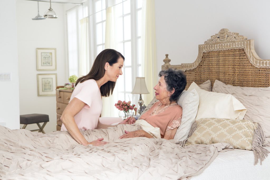 A caregiver and an elderly woman share a moment in a cozy bedroom setting.