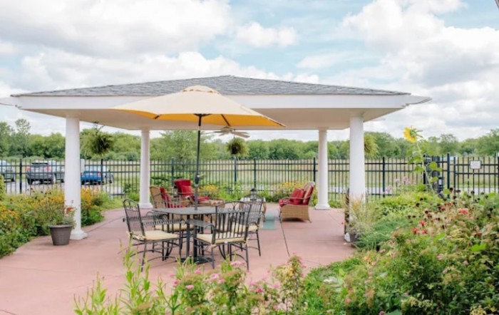 A tranquil outdoor patio area featuring a table with chairs and a large umbrella under a gazebo in a garden setting.