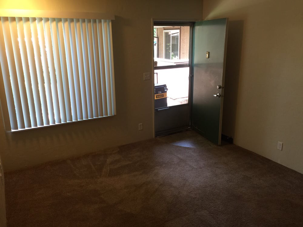 A small empty room with carpet flooring, a window with vertical blinds, and an open door leading outside.