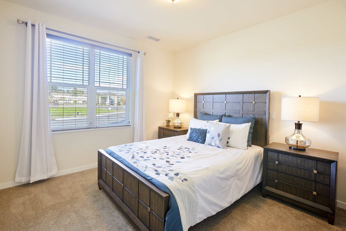 A well-lit bedroom featuring a neatly made bed with matching nightstands and lamps, complemented by a large window allowing natural light.
