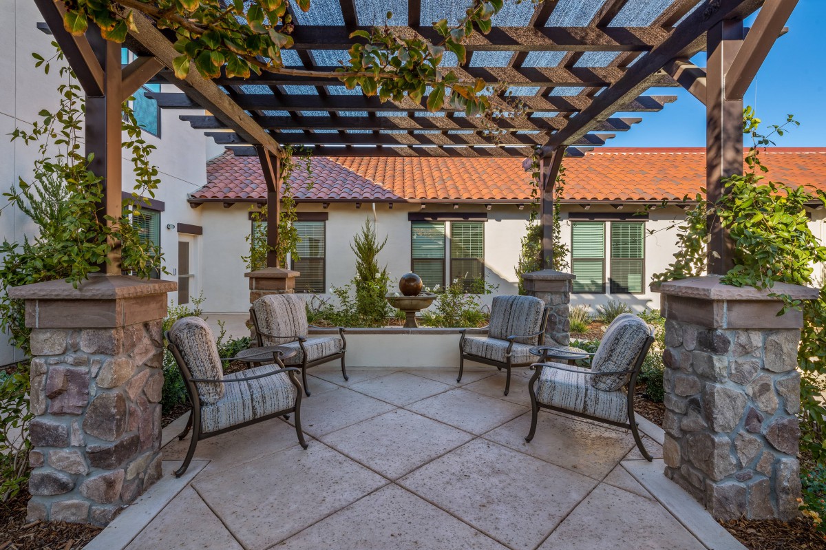 A serene courtyard with comfortable chairs under a pergola, featuring a central fountain and surrounded by greenery.