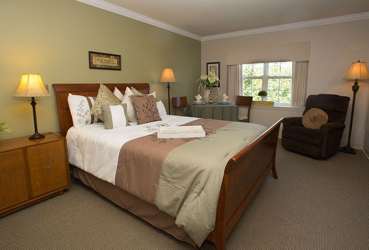 A well-decorated hotel room featuring a large bed with pillows and throws, bedside lamps, and a window overlooking greenery.