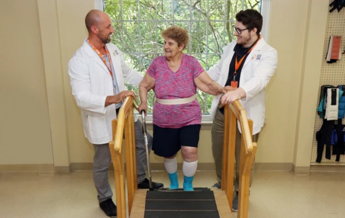 An older adult receives assistance from two healthcare professionals during a physical therapy session using exercise equipment.