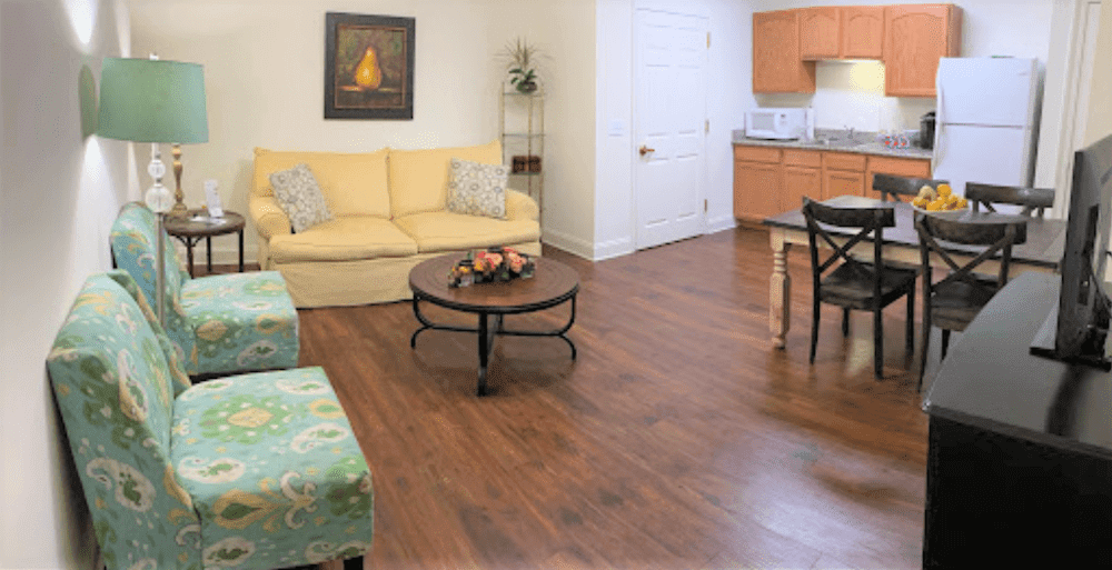A cozy living room with a yellow sofa, wooden flooring, and a connected kitchen area.