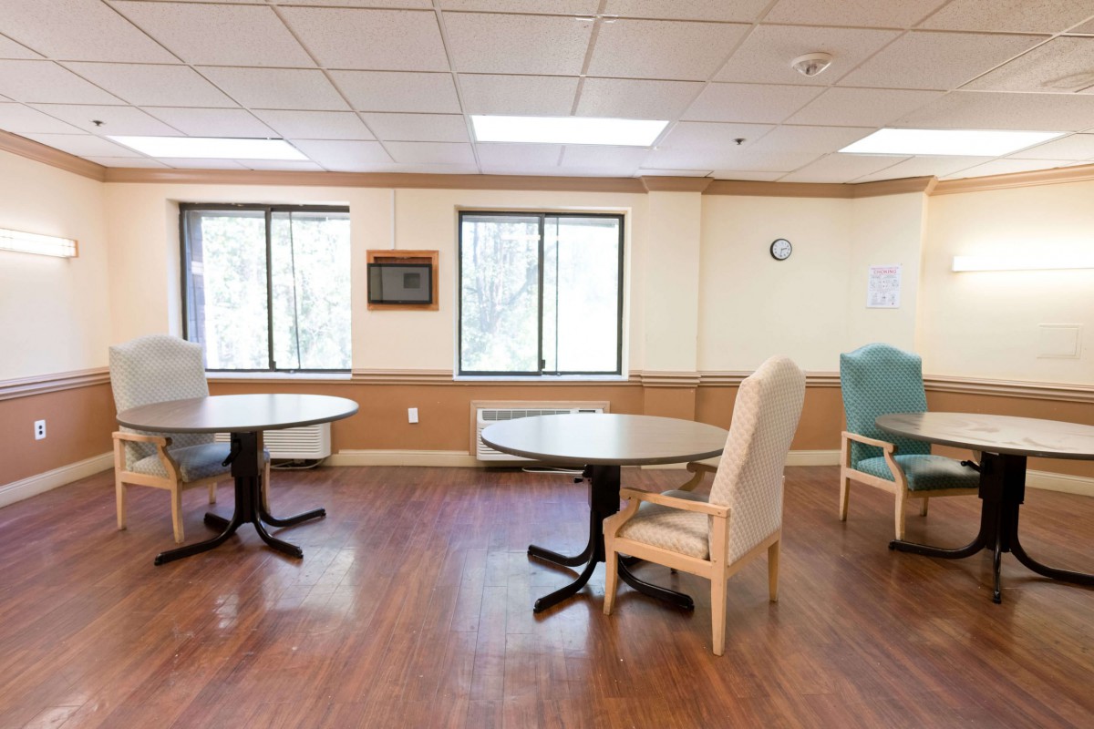 An empty room with wooden floors, tables, and chairs near large windows.