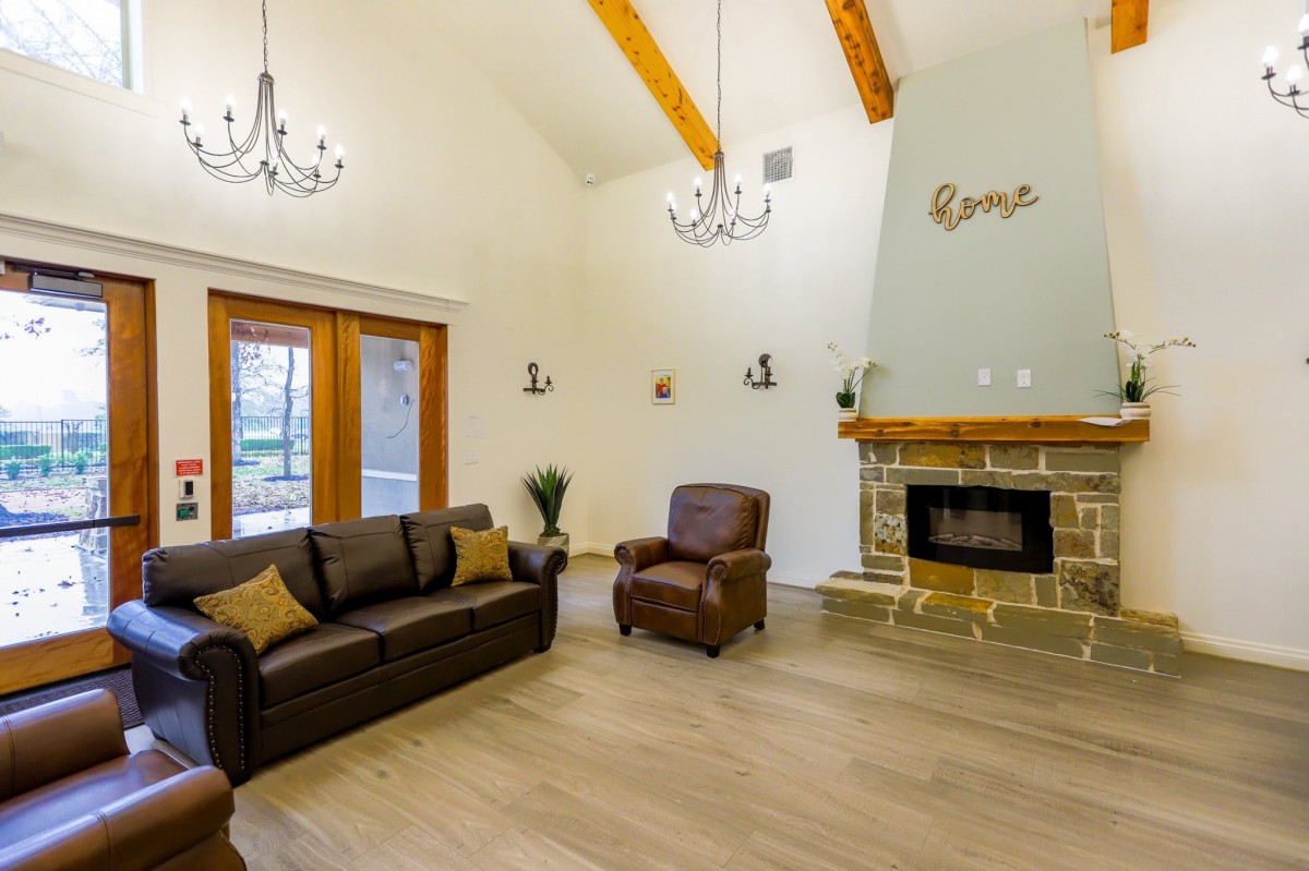 A spacious living room featuring a cozy sofa and armchair, a stone fireplace, and elegant chandeliers under wooden ceiling beams.
