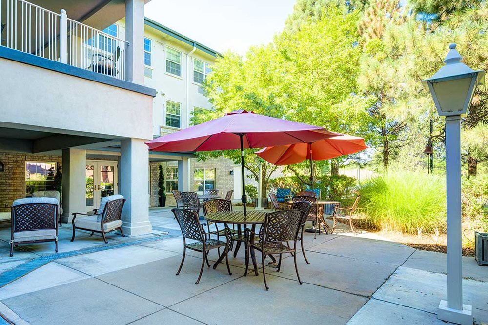 A sunny patio area with a table and chairs under a large red umbrella, surrounded by greenery and adjacent to a building.