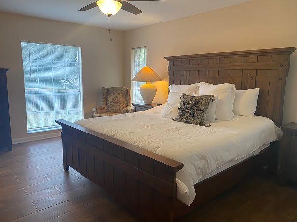 A cozy bedroom featuring a large bed with multiple pillows, a lamp on the side table, and natural light coming through the windows.