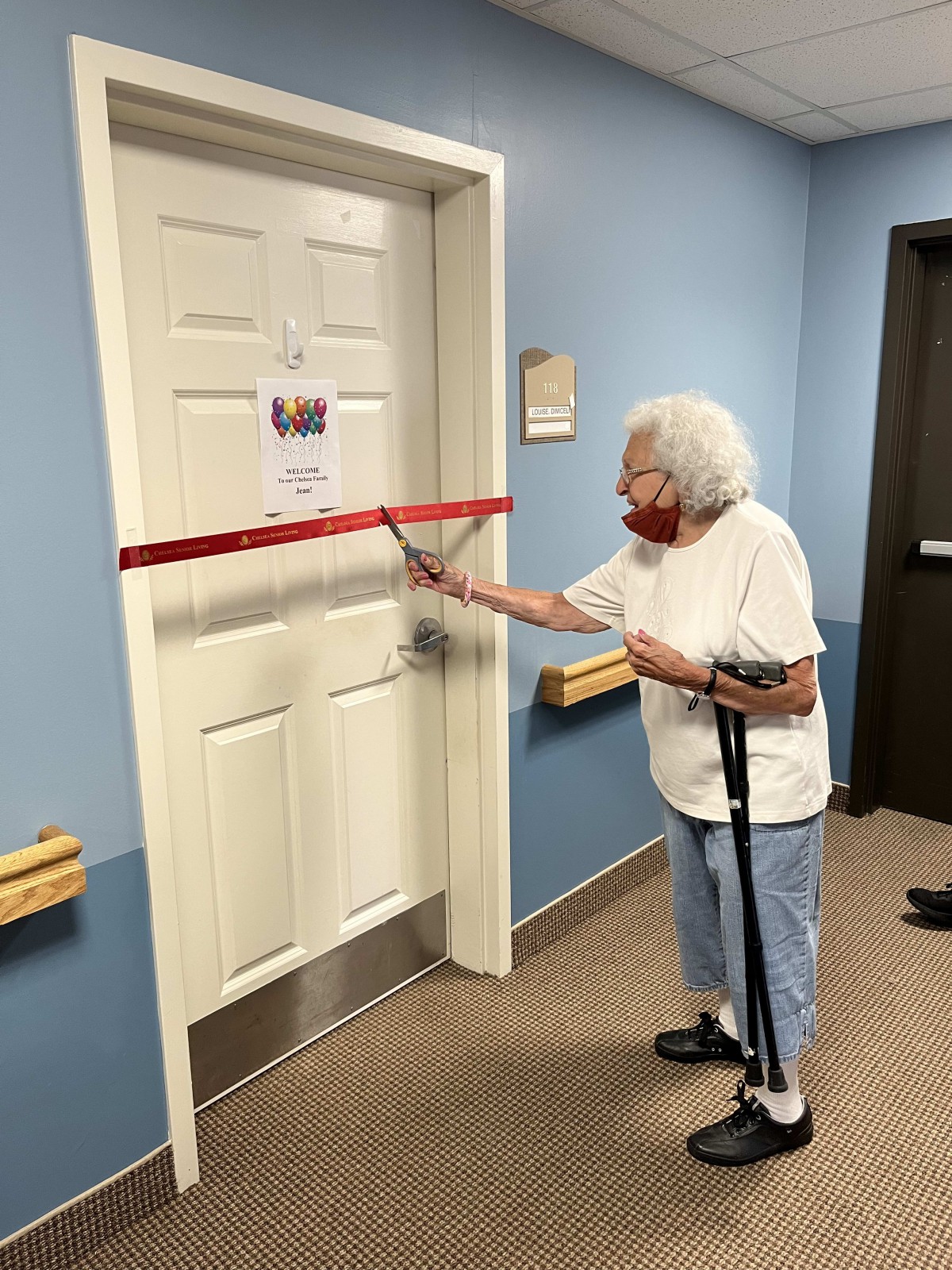 An elderly woman using scissors to cut a ribbon in front of a door, marking a ceremonial occasion.