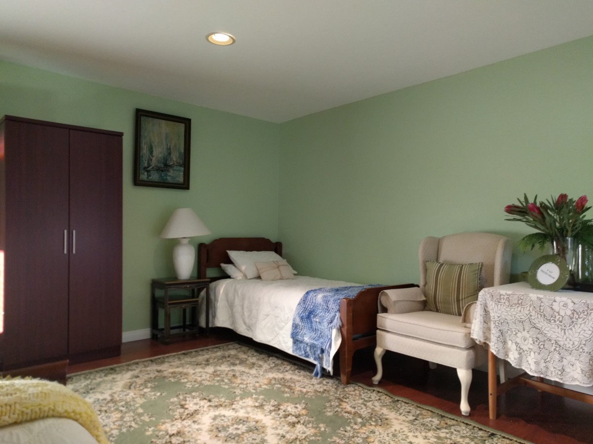 A cozy bedroom with green walls, featuring a single bed, an upholstered chair, and a side table with a lamp. A piece of artwork hangs on the wall, and an ornate rug covers the floor.
