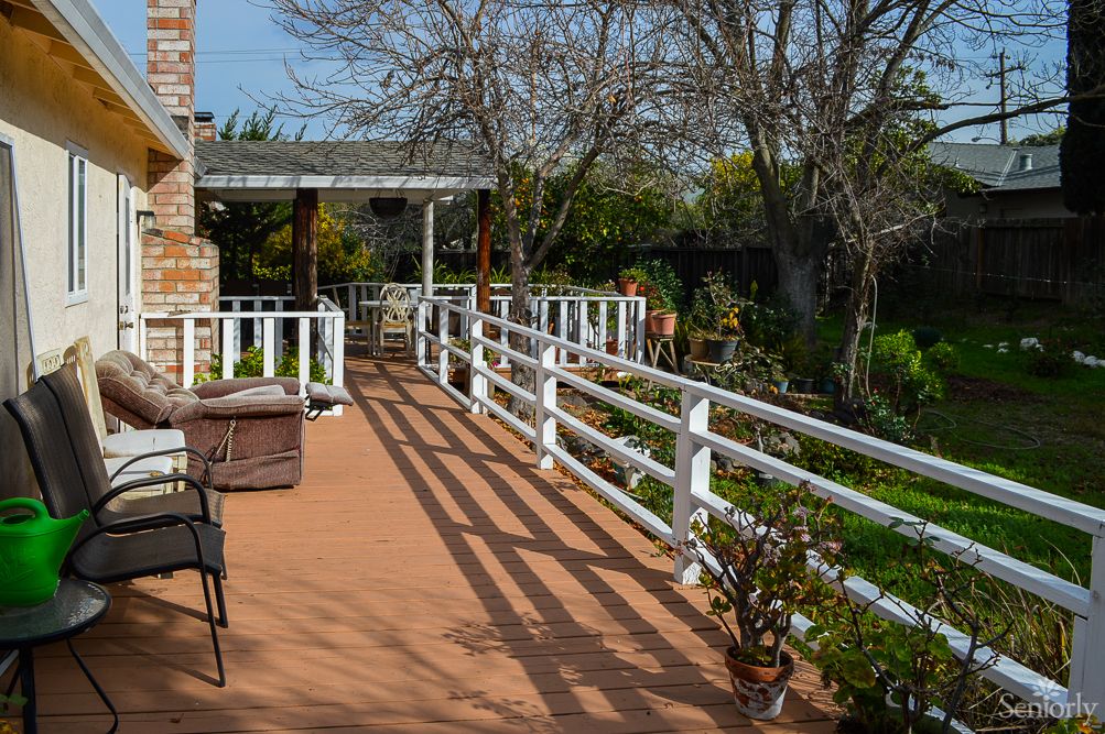 A sunny outdoor deck overlooking a garden, surrounded by a white fence and featuring patio furniture.