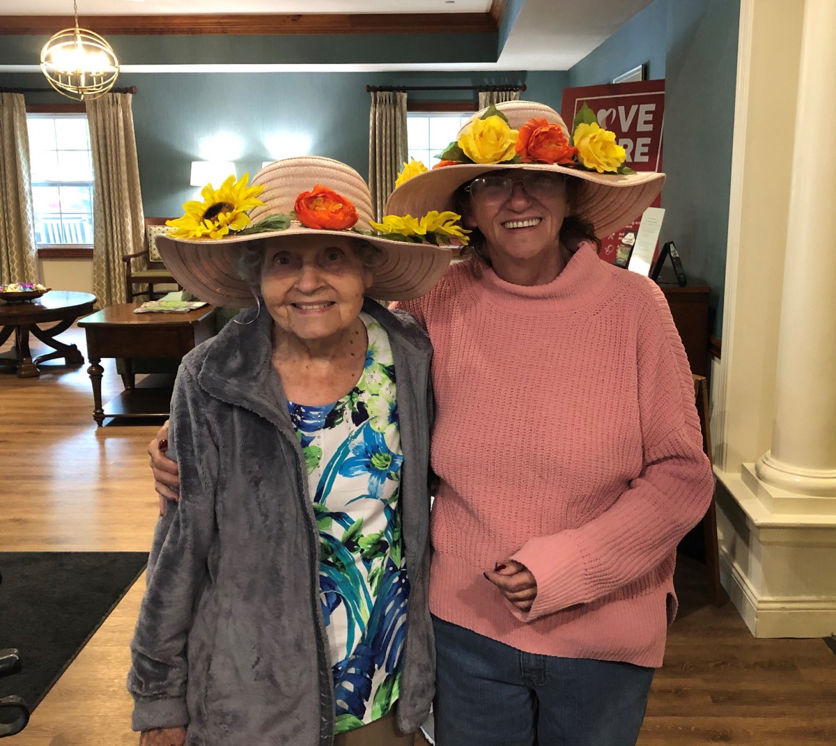 Two women smiling and wearing colorful hats adorned with flowers.