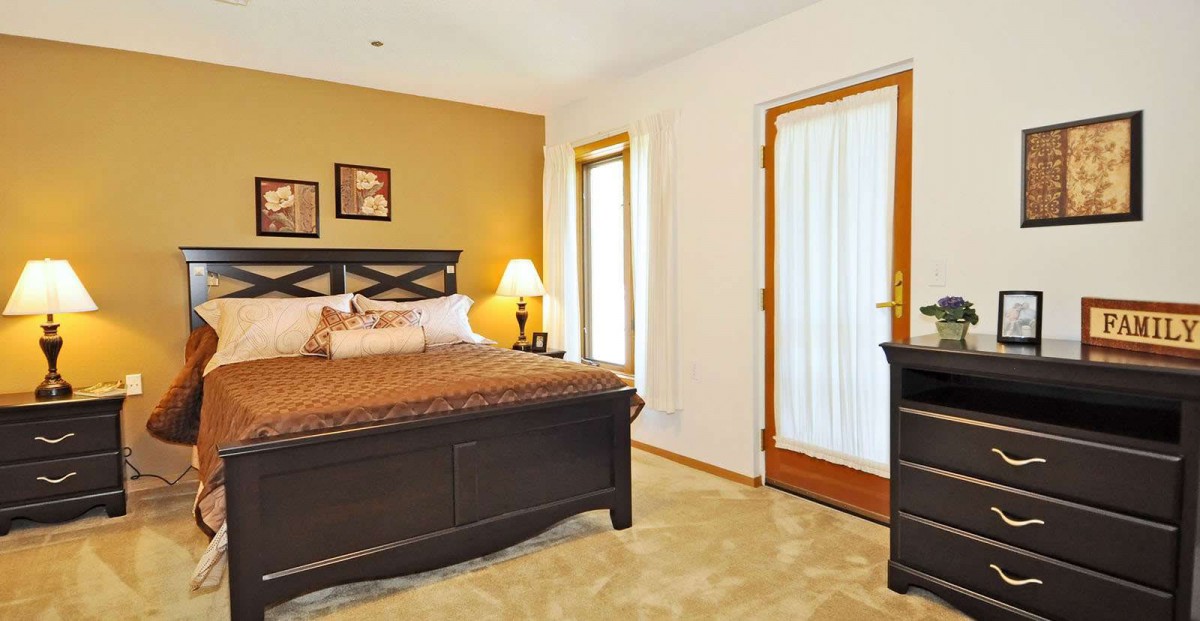 A cozy bedroom with a comfortable bed, warm lighting from a lamp, and natural light streaming through the windows.