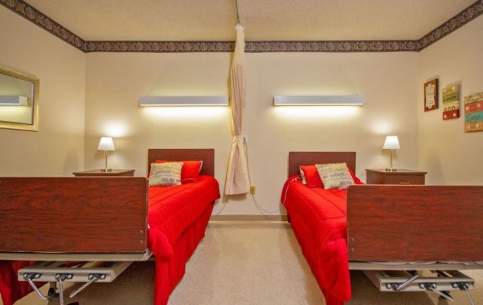 A hospital room with two twin beds featuring red bedding and warm lighting.