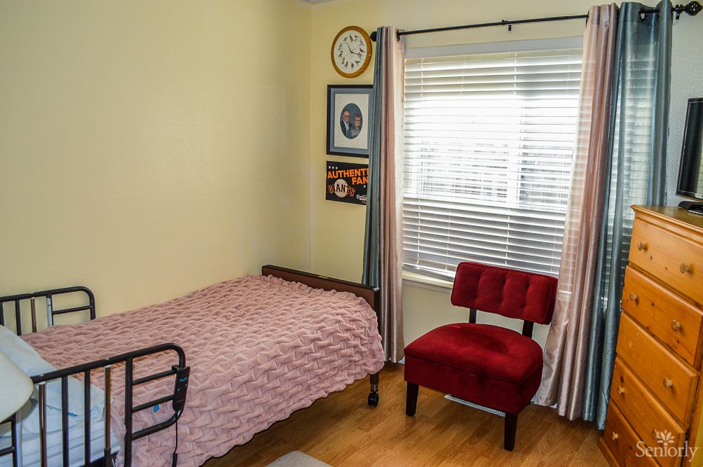 A cozy bedroom with a single bed, red chair, and light streaming in through a window.