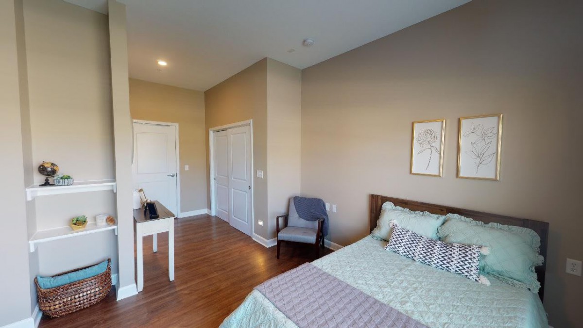 A cozy bedroom with a neatly made bed, wooden flooring, a chair, and minimalistic wall decor.