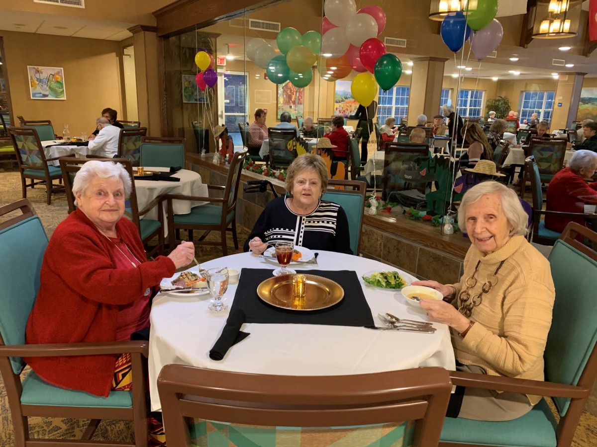 Three elderly women enjoying a meal together at a decorated dining area with colorful balloons.