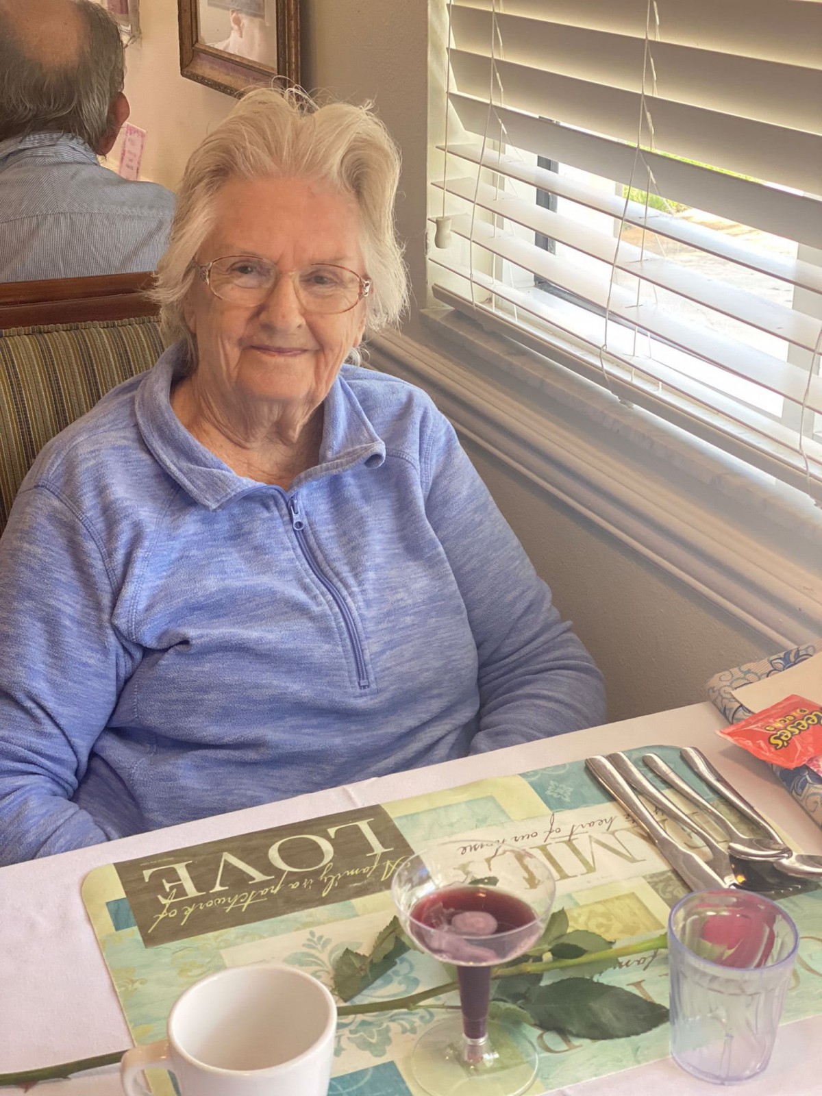 An elderly woman sitting at a table in a restaurant, smiling with daylight shining through the window.