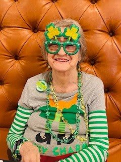 An elderly woman celebrates St. Patrick's Day with festive green accessories and shamrock-themed glasses.