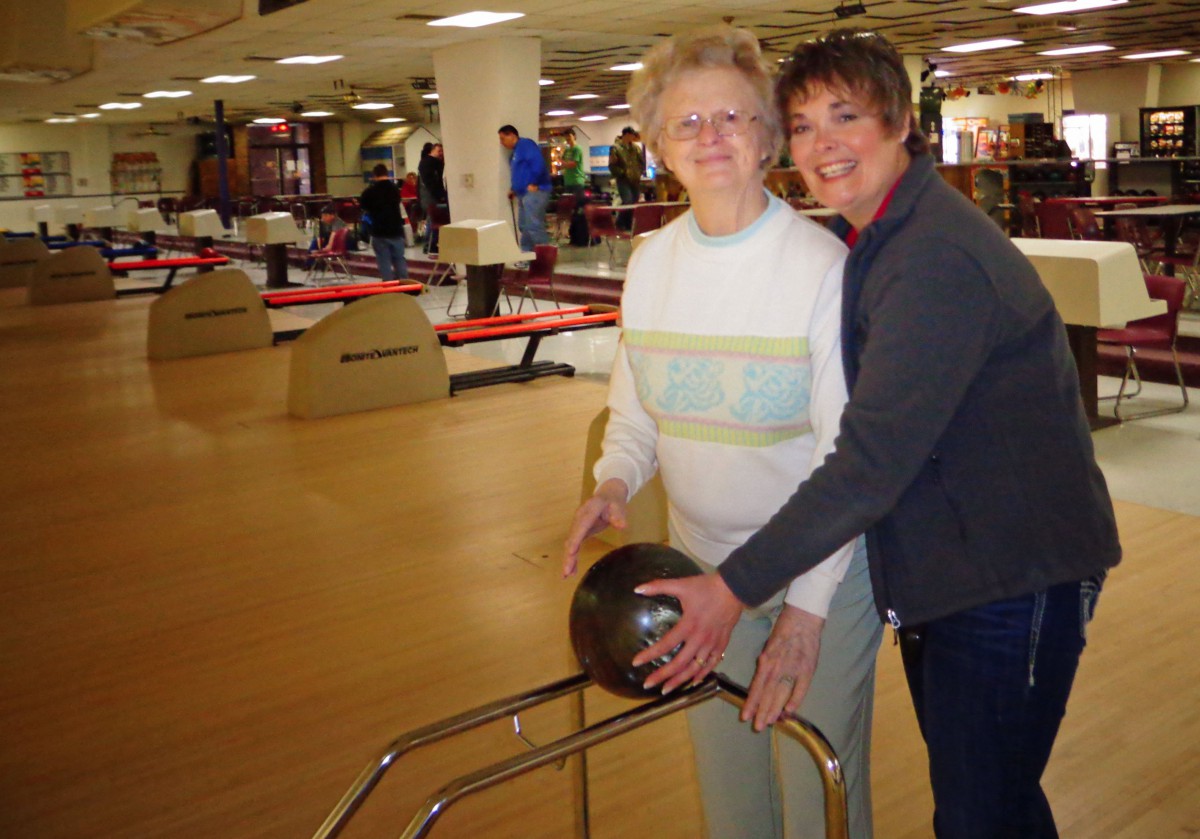 An elderly woman receiving assistance while bowling in an indoor sports facility.
