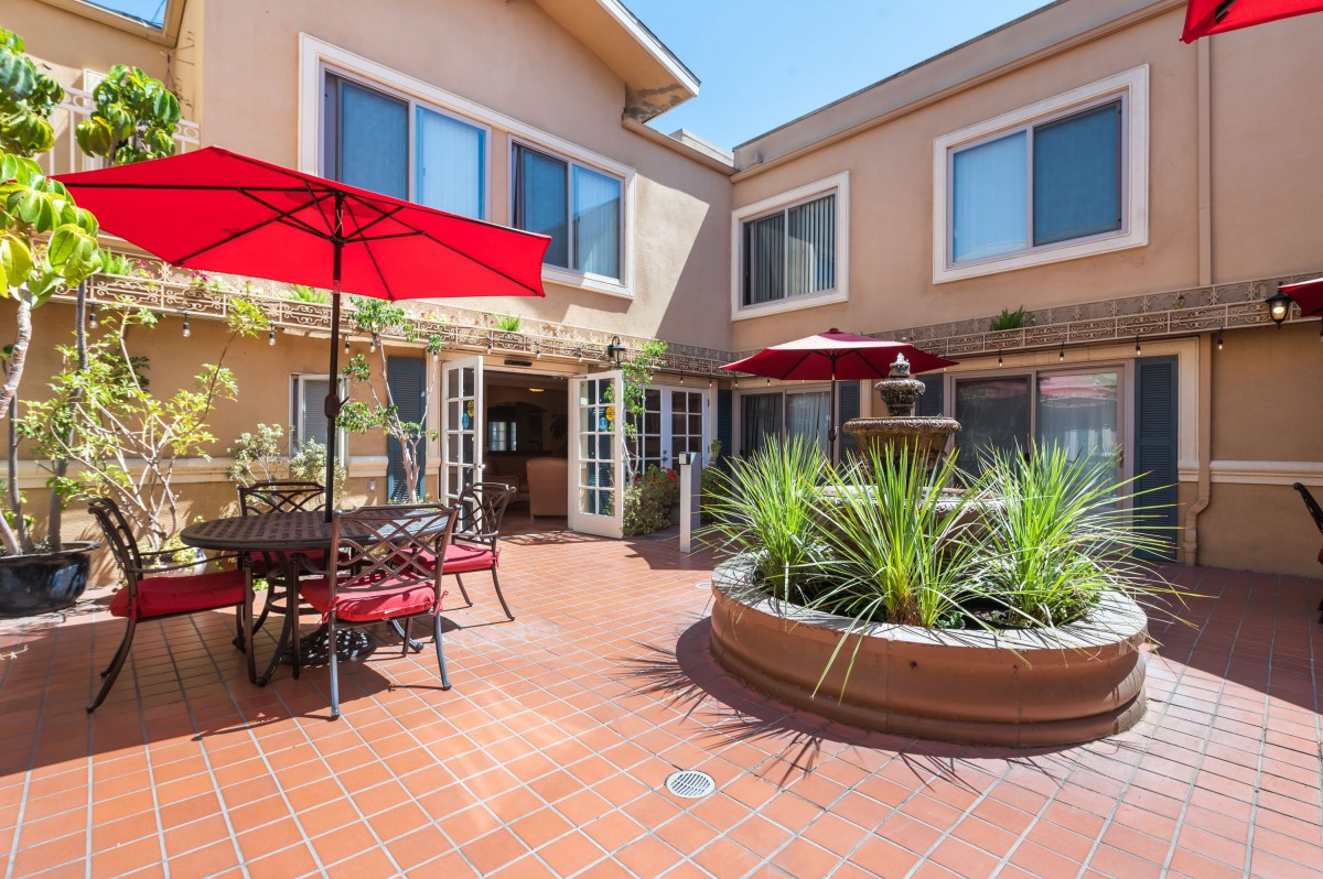 A sunny courtyard with patio furniture and red umbrellas, surrounded by windows.