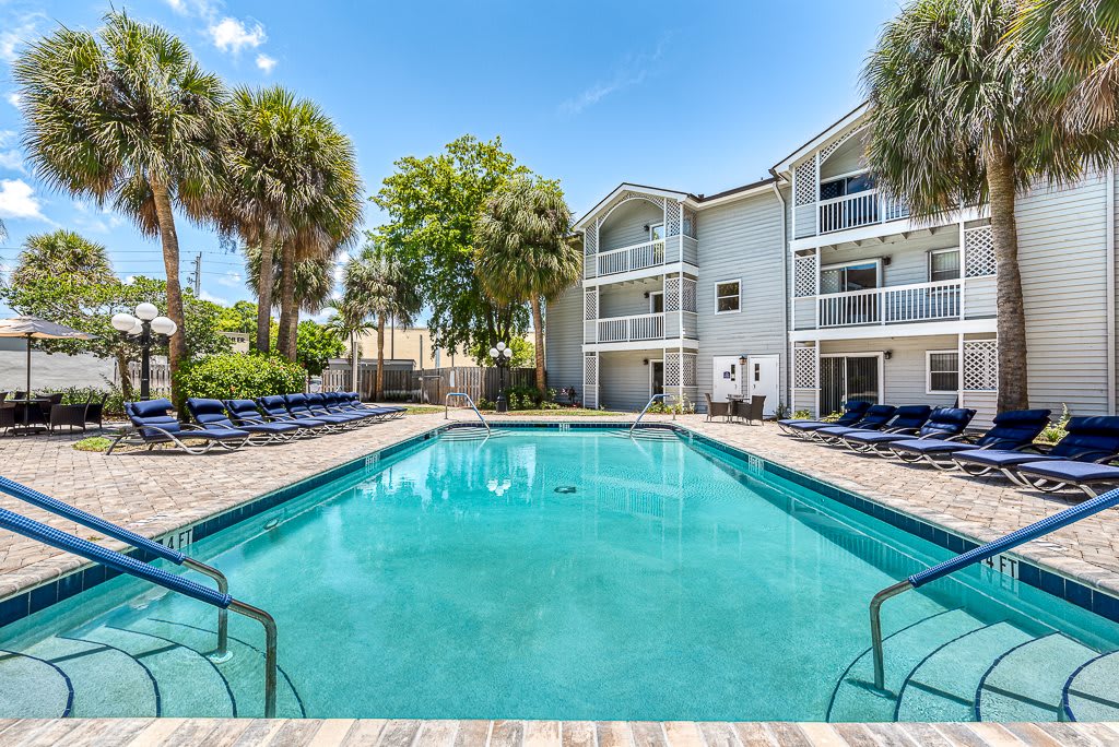 A serene outdoor swimming pool surrounded by apartment buildings and palm trees under a clear, sunny sky.