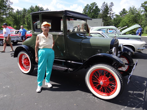 An elderly individual proudly stands next to a vintage green car with striking red wheels at a car show.
