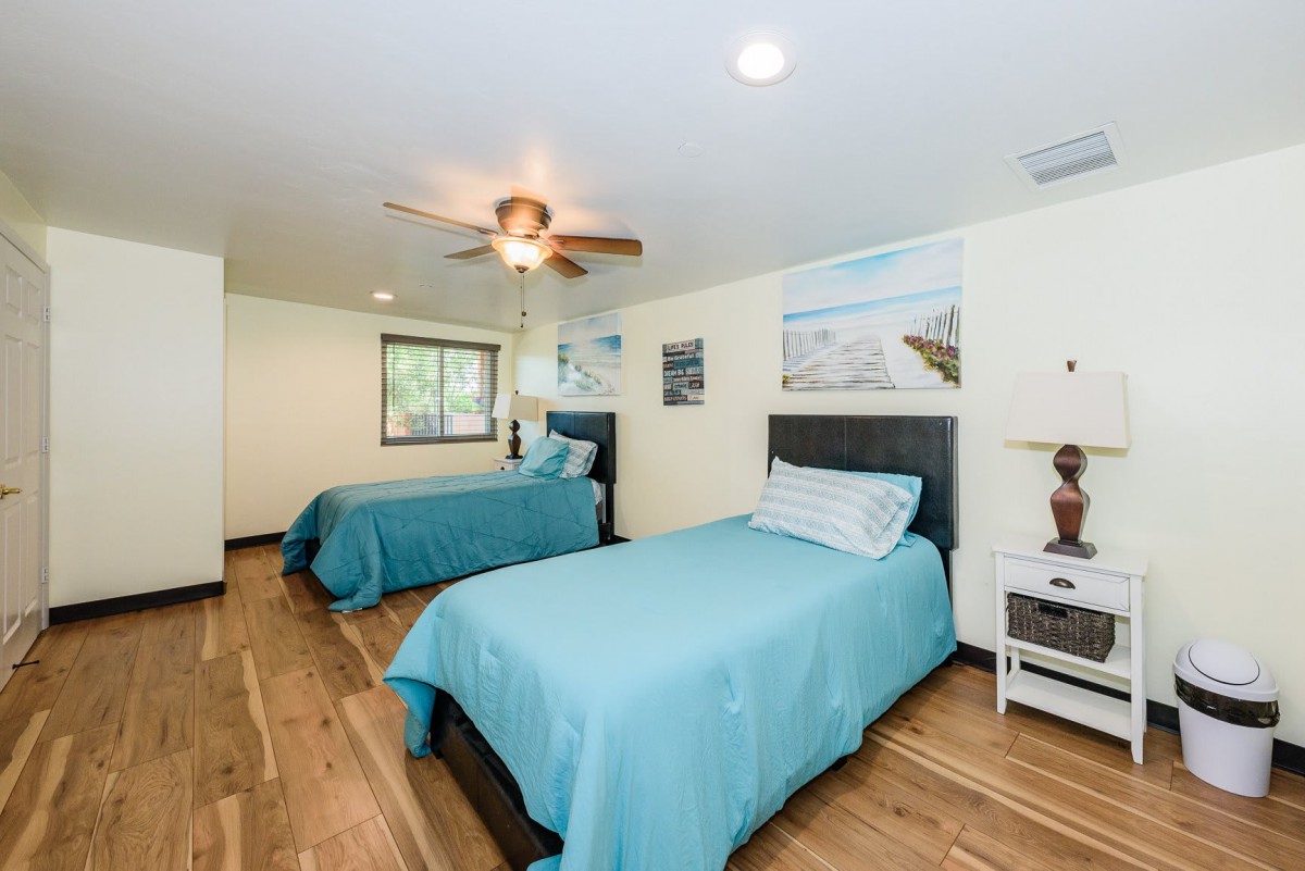 A modern bedroom with two twin beds and blue bedding, featuring a ceiling fan and wood flooring.