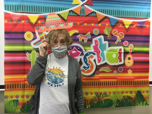 A person wearing a vibrant sombrero and a mask poses joyfully in front of a colorful festive backdrop.