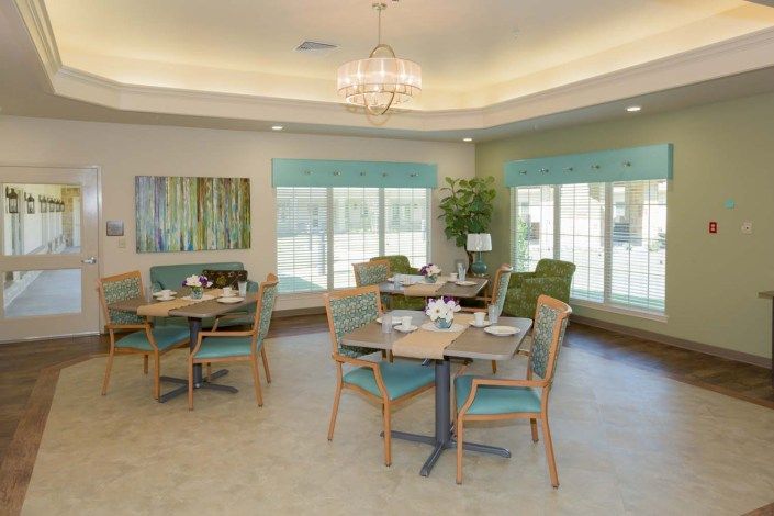 A bright and elegantly designed dining room with multiple tables and chairs, featuring large windows and a chandelier.